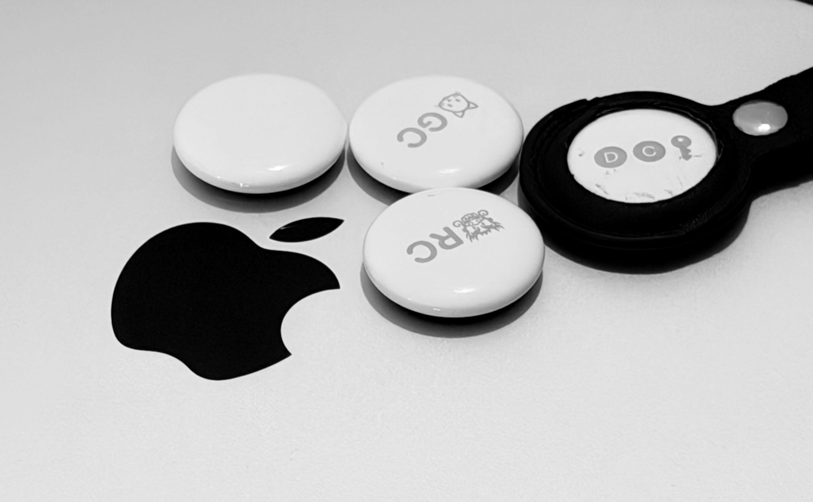 A black and white photograph of four AirTags resting on the lid of a MacBook, beside the engraved Apple logo. Three lie loose and one sits in a black keyring holder. Each is engraved with initials and a small emoji.