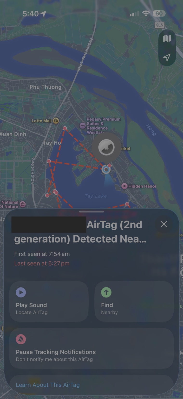 An iPhone Find My screen. A dark satellite map of Tay Ho, Hanoi, with a red dashed line tracing a route around West Lake and a grey AirTag marker over the water. A panel below reads 'AirTag (2nd generation) Detected Nea...', with the owner's name covered by a solid black redaction box, then 'First seen at 7:54 am' and, in red, 'Last seen at 5:27 pm', above Play Sound, Find and Pause Tracking Notifications buttons.